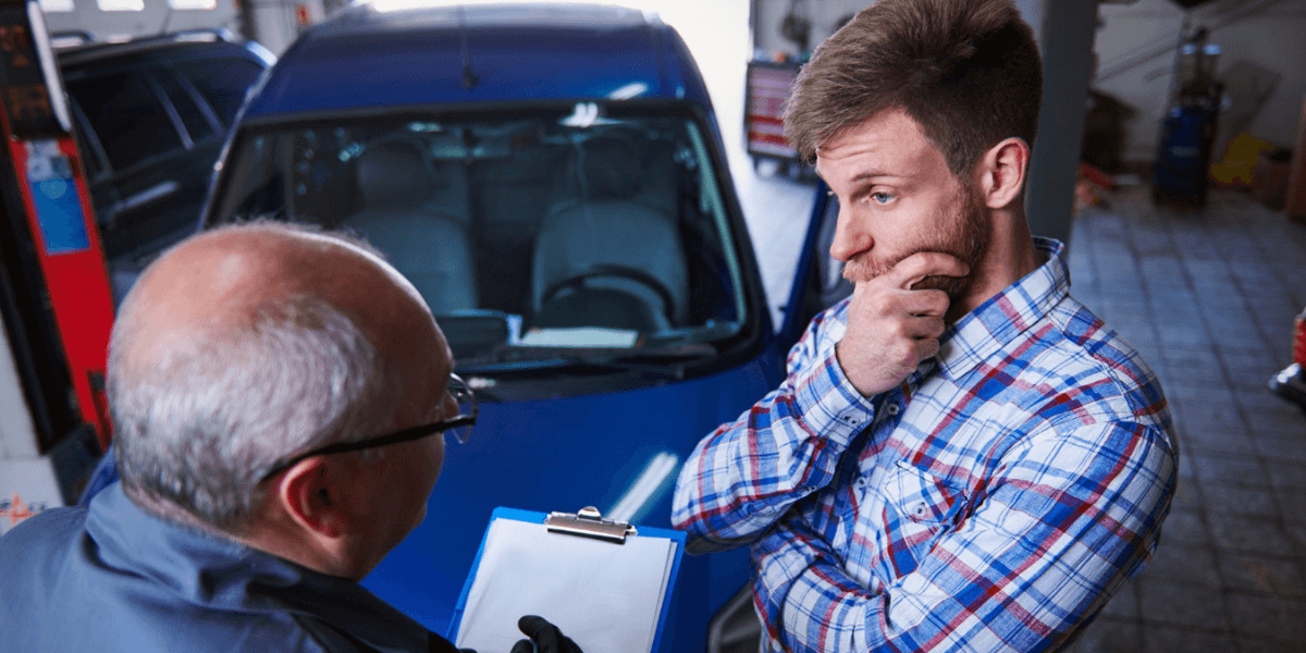 A man converses with another man inside a car repair shop, surrounded by tools and vehicle parts.